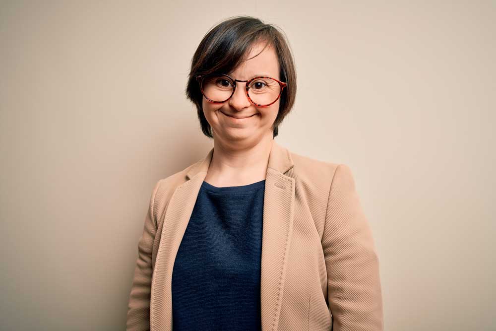 junge Geschäftsfrau mit kurzen dunkelbraunen Haaren mit Down-Syndrom lächelt. Sie trägt eine braune runde Brille, einen hellen Blazer und ein blaues Shirt.